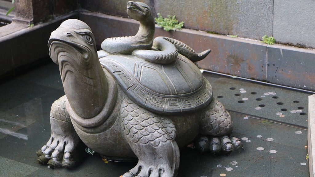 A stone statue of a turtle with a snake coiled on its back, partially submerged in water. The turtle has a fierce expression and detailed carvings on its shell and limbs.