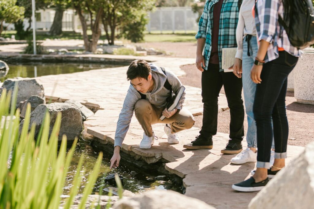 Three young people gaze thoughtfully at a serene pond, surrounded by lush greenery and soft sunlight filtering through trees.