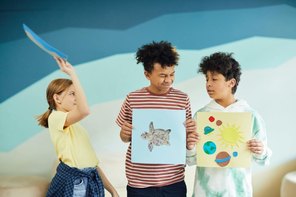 Three children proudly display their colorful drawings in a bright, cheerful room filled with art supplies.